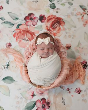 Newborn wrapped in white blanket sleeps on pink cushion with floral backdrop featuring coral roses and greenery.
