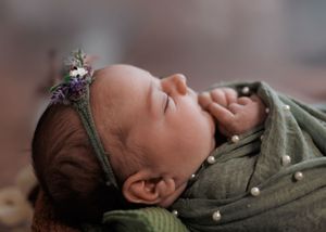 A sleeping infant wearing olive green clothing and a delicate floral headband rests peacefully.
