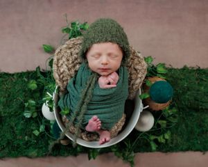 A newborn sleeping peacefully in a rustic bowl surrounded by greenery and wearing a sage green wrap and knitted hat.