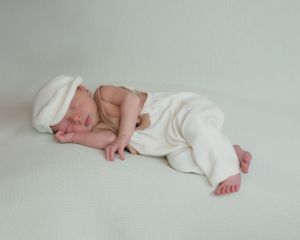 A sleeping newborn baby in cream colored wrap lies curled up on a light colored backdrop.