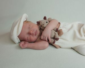 A sleeping baby in white clothing cuddles with a brown teddy bear on a light-colored backdrop.