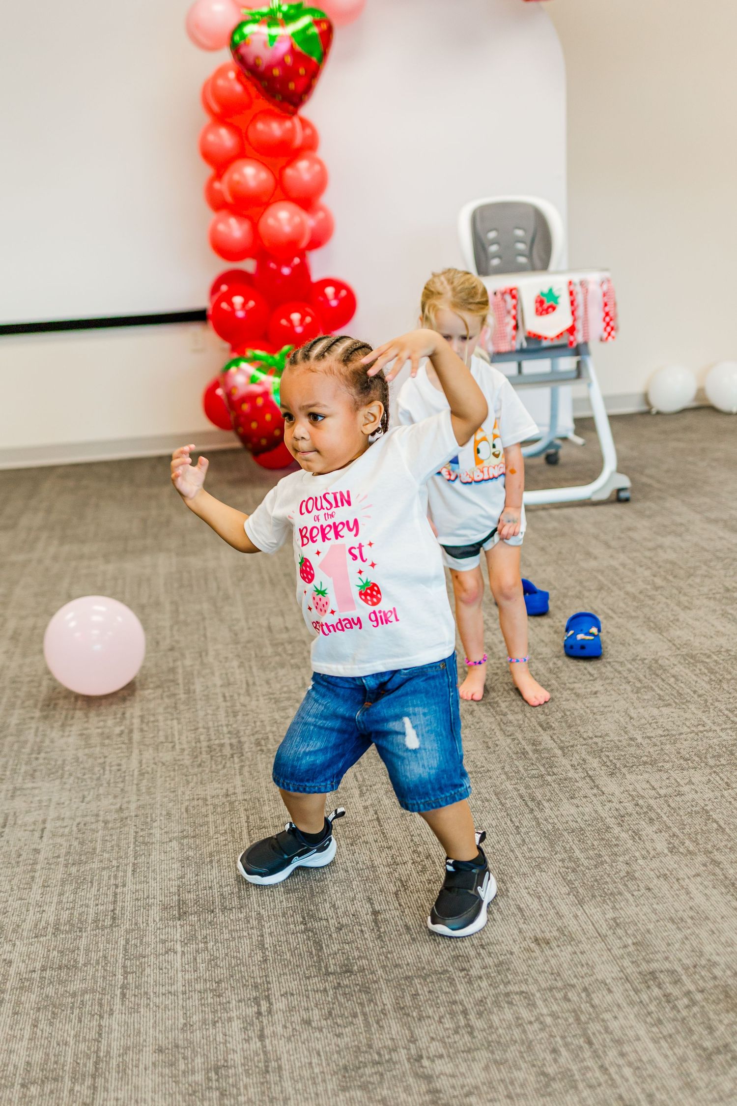 A child in a berry-themed shirt dances near red balloon decorations at a party.