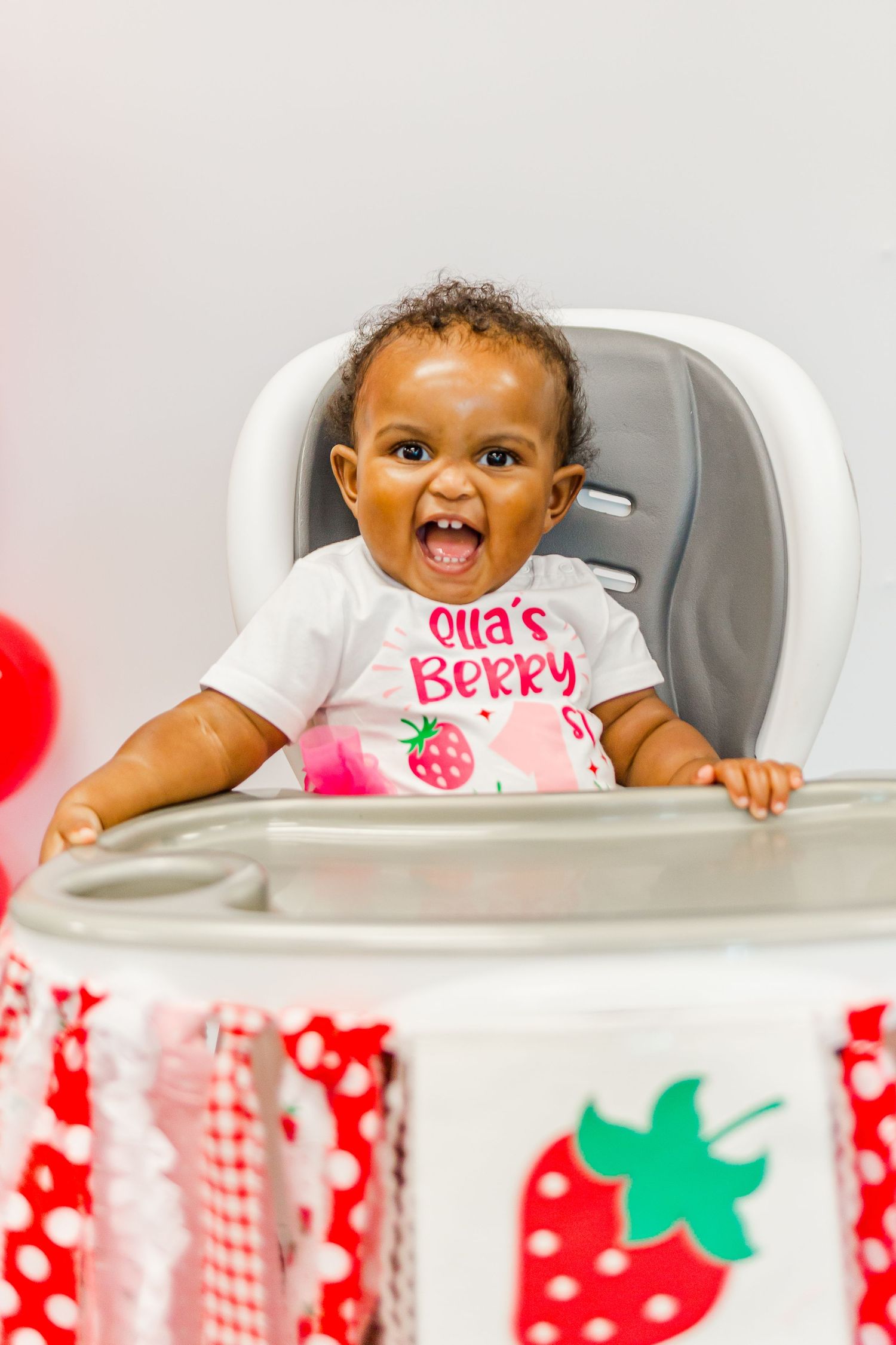 Baby wearing a strawberry-themed bib sits smiling in a high chair.