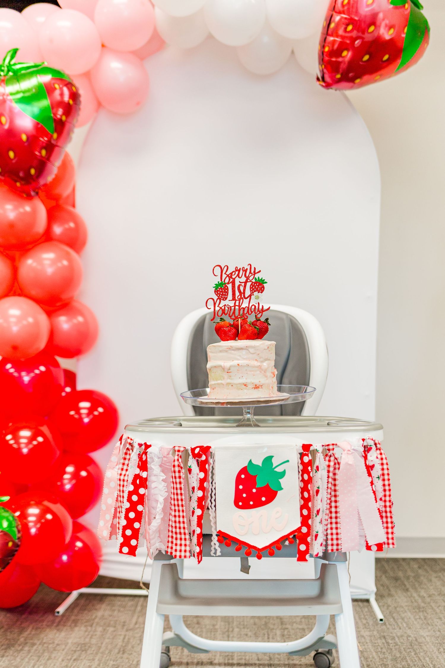 Strawberry themed birthday party setup with balloon arch and high chair decorated with red fabric.