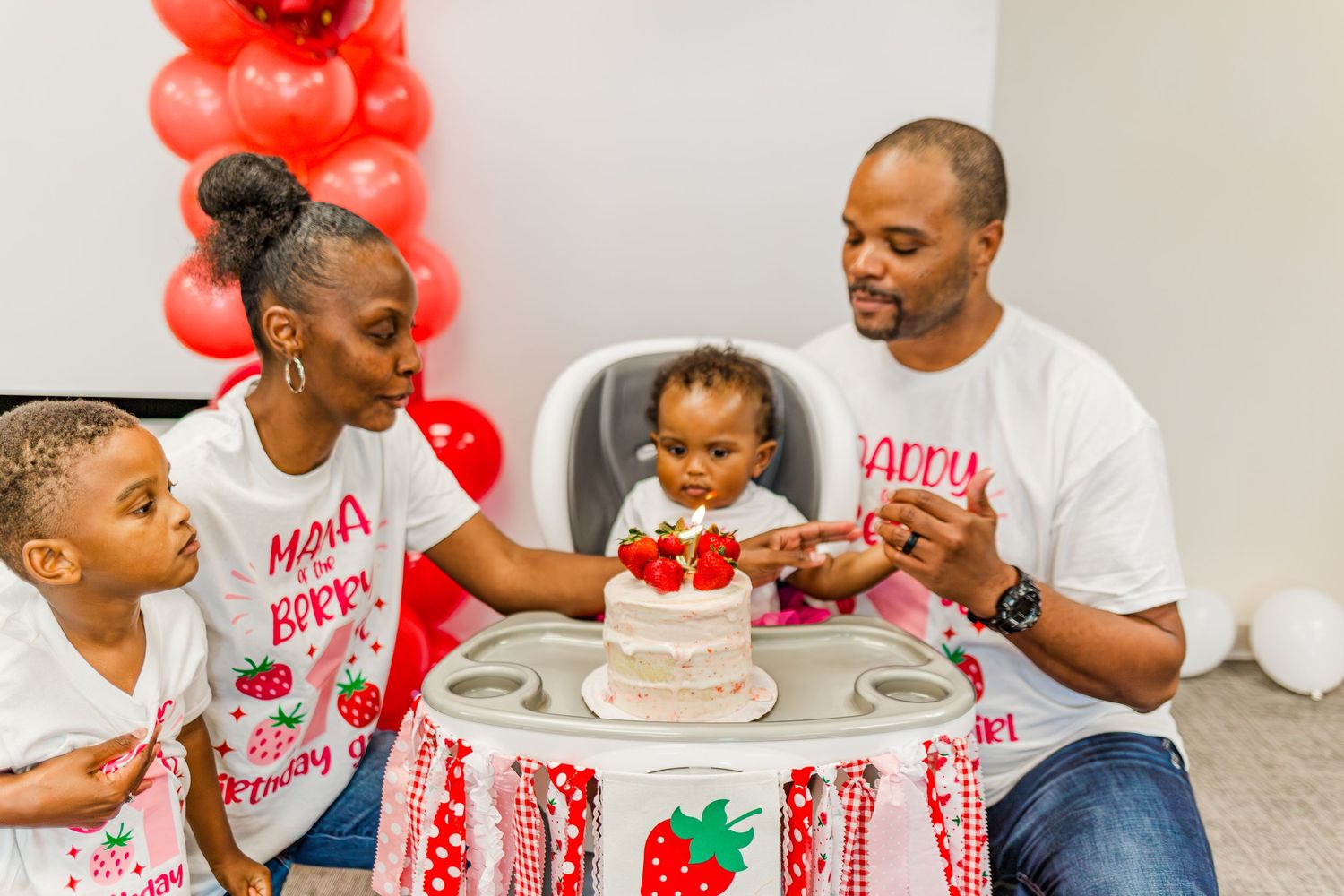 Family gathered around a decorated high chair for a strawberry-themed celebration.