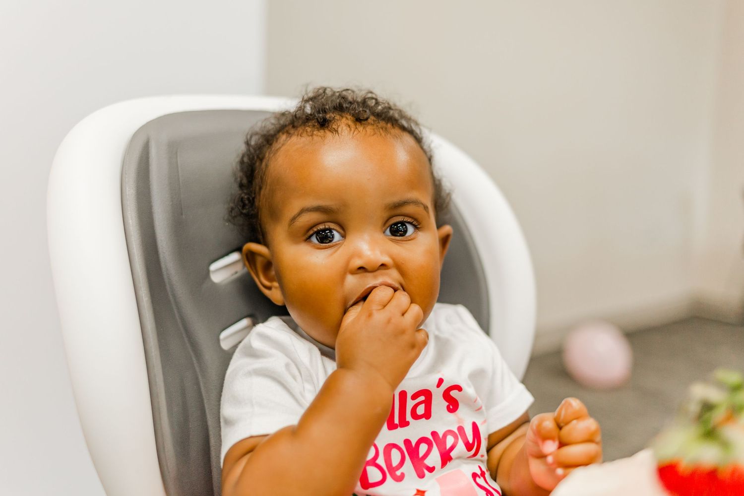 Young child in white highchair with finger in mouth during a celebration.