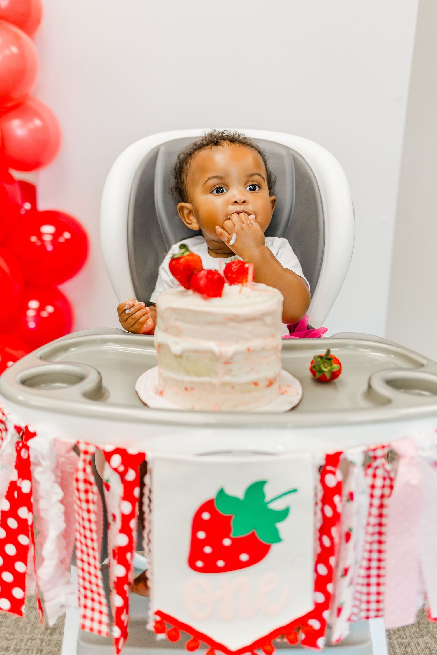 Baby in decorated high chair with strawberry themed cake and red balloon decorations.