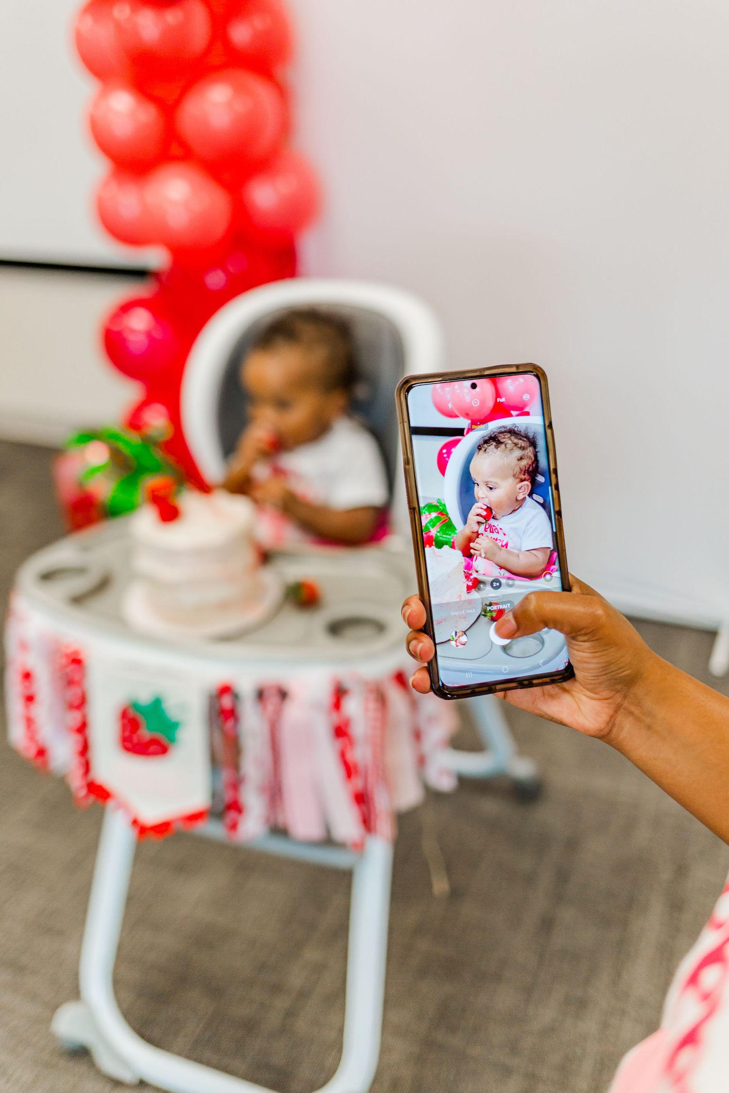 Hand holding phone to capture photo of baby enjoying cake in decorated high chair.
