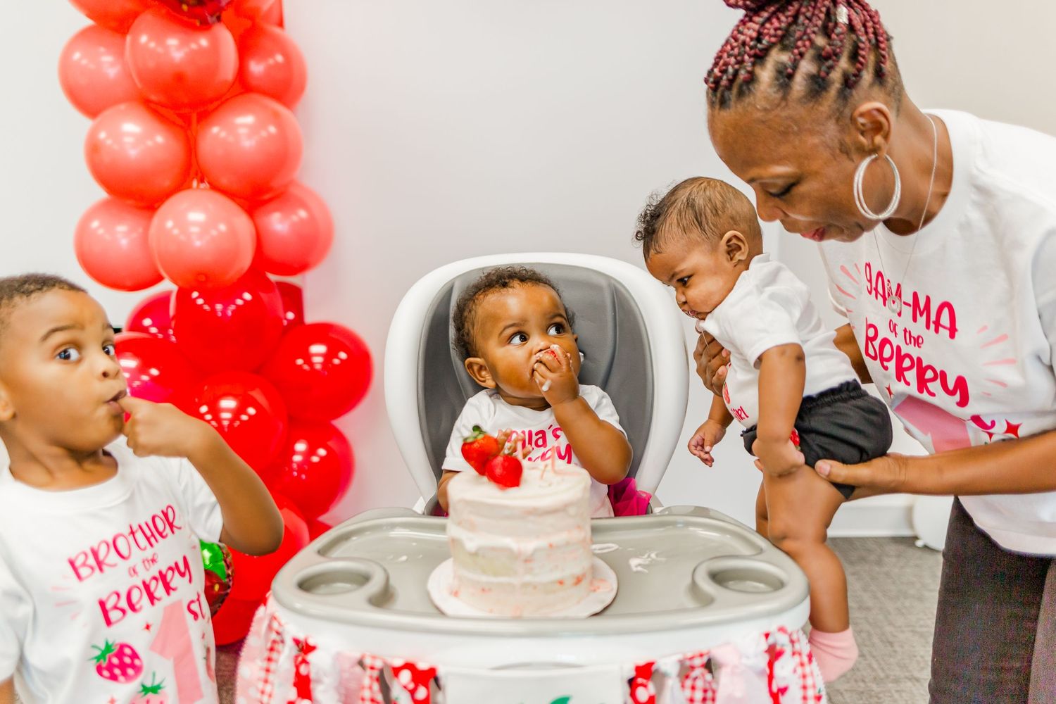 A festive birthday celebration with a white cake surrounded by red balloon decorations and strawberry themed decor.