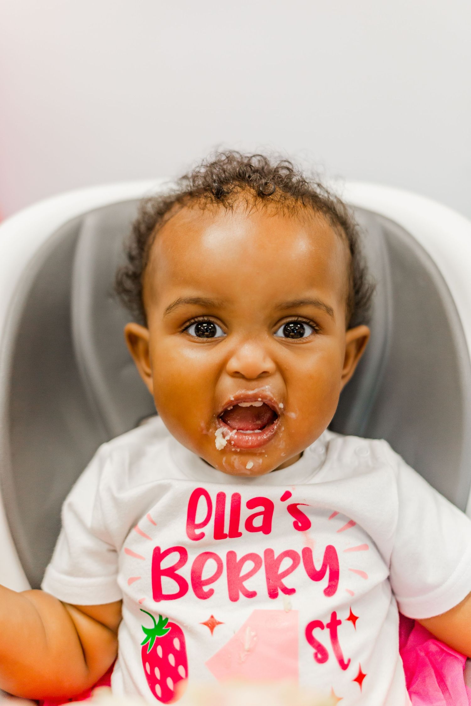 A close up shot of a toddler in a white strawberry-themed t-shirt smiling with excitement.