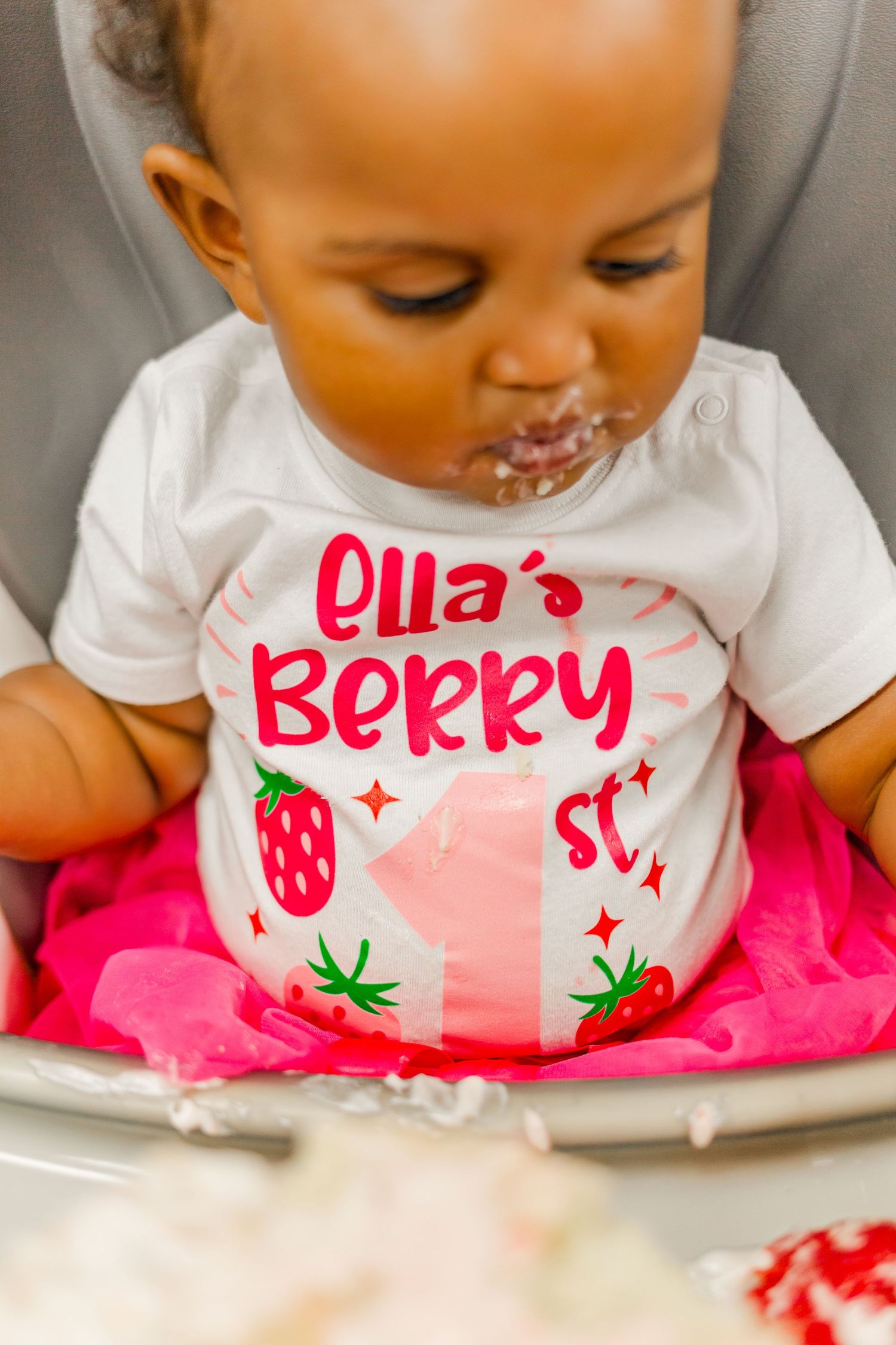 A baby wearing a strawberry-themed shirt with cake frosting on their face.