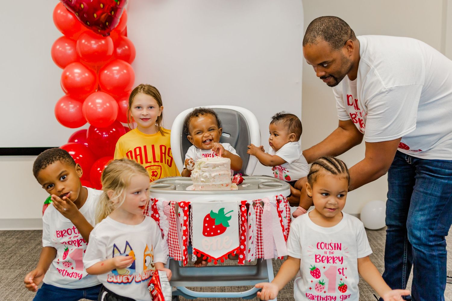 A group gathering around a birthday cake with strawberry-themed decorations and red balloons.