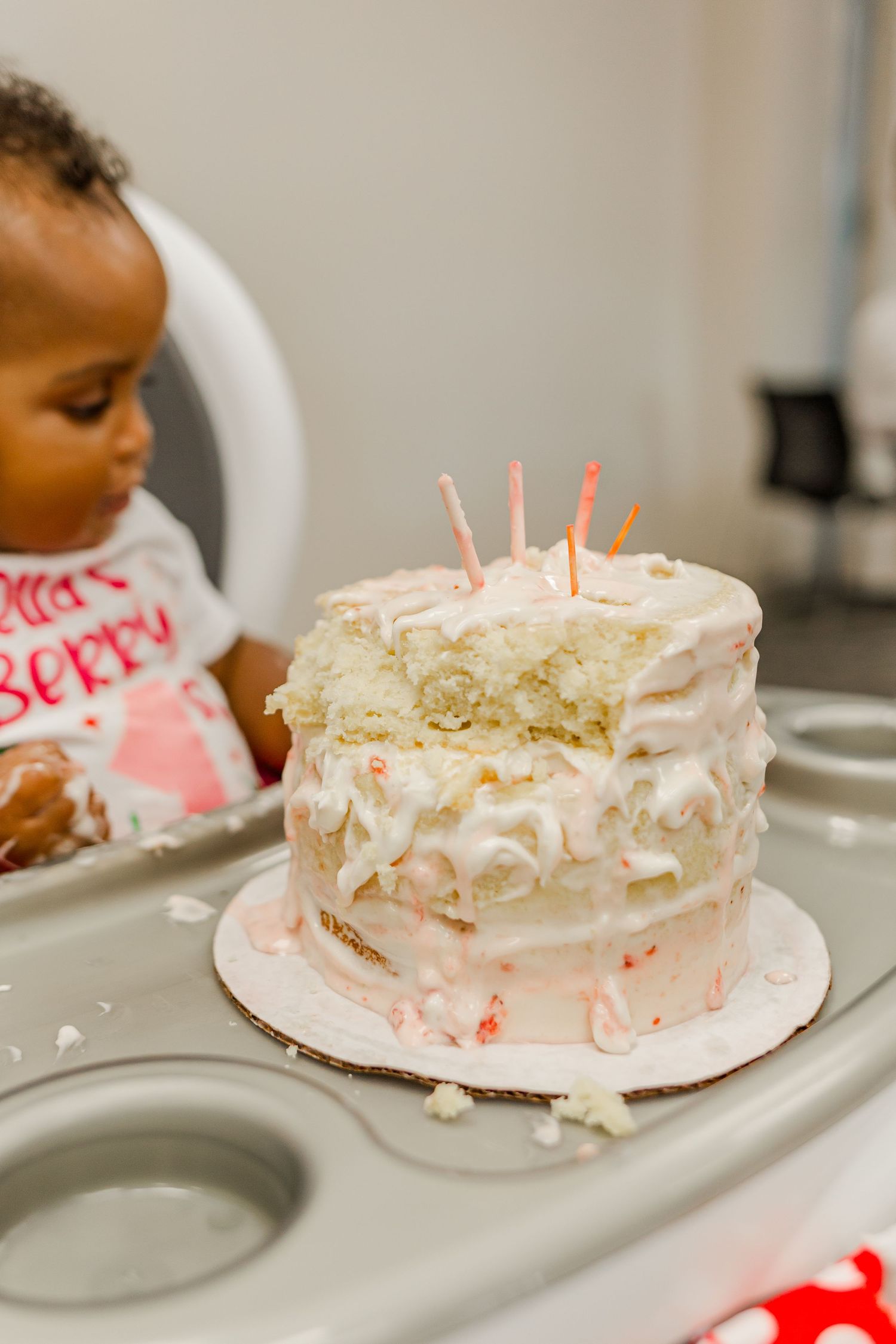 A white birthday cake with pink candles sits on a high chair tray during a celebration.