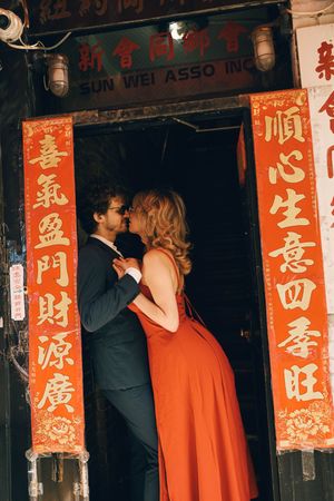 Couple sharing a romantic kiss in a red dress and suit framed by traditional Chinese red banners in a doorway.