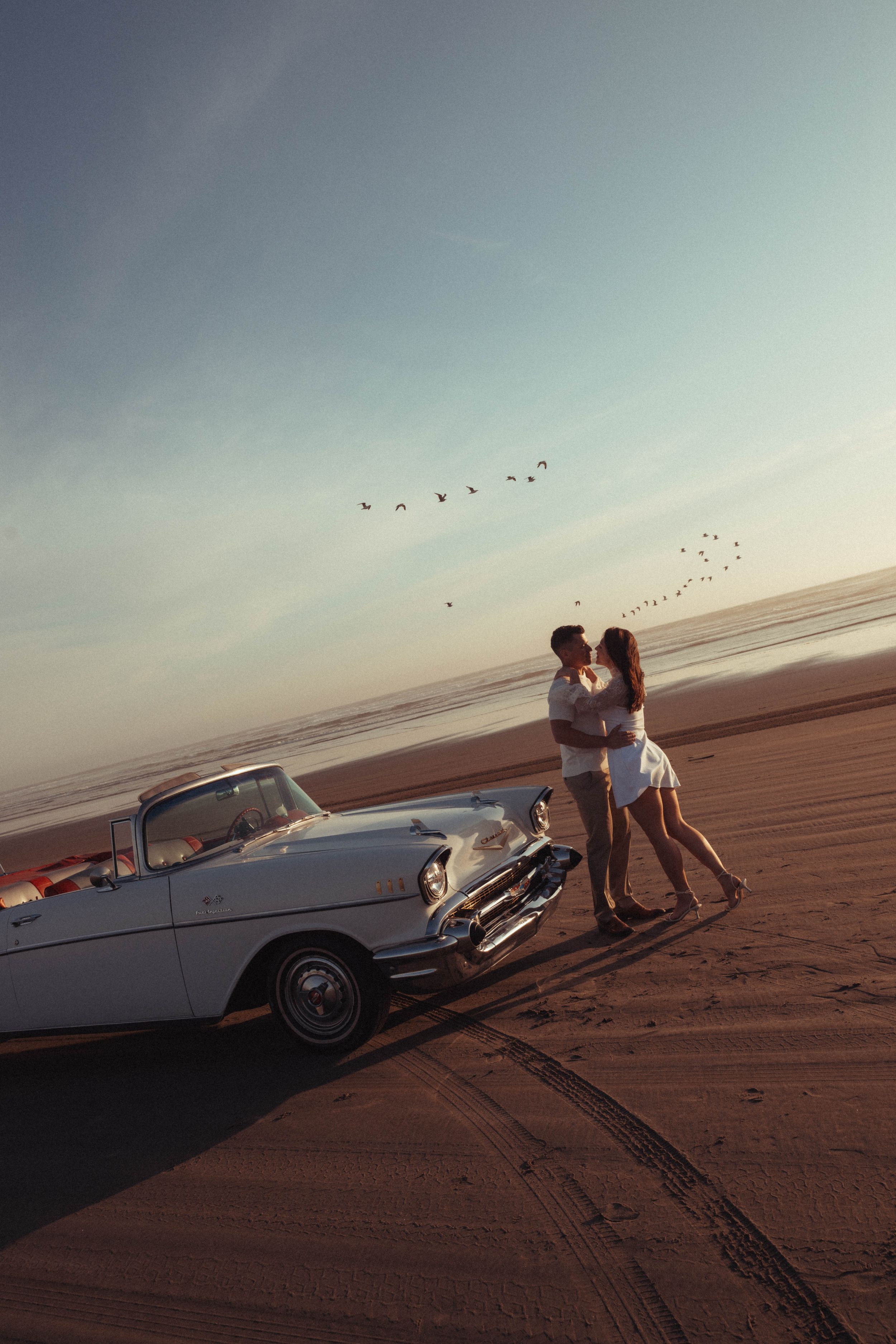 Couple kissing beside a classic white convertible car on a sandy beach at sunset with birds flying overhead.