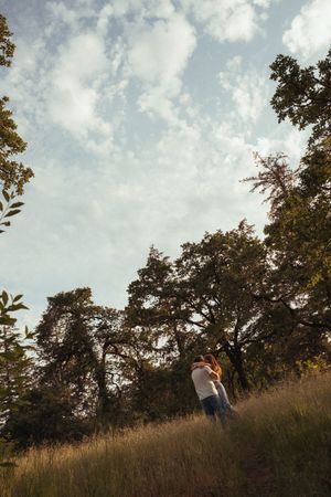 Couple embracing on a grassy hillside surrounded by tall trees under a partly cloudy sky.