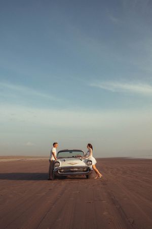 Couple standing by a classic vintage convertible car on a sandy beach under a blue sky at sunset.