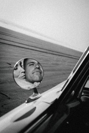 Black and white photo of a man and woman reflected in a vintage car side mirror on an open road.