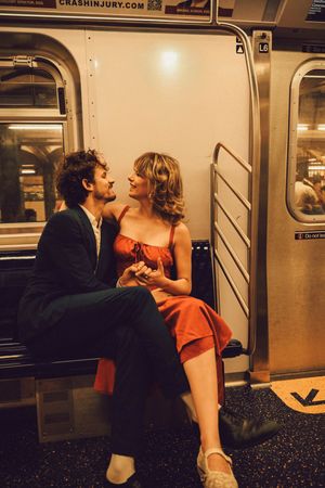 Couple holding hands and smiling on a subway train, woman in red dress seated on man's lap.