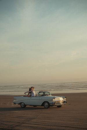 Couple embracing beside a classic vintage convertible car parked on a sandy beach at sunset.