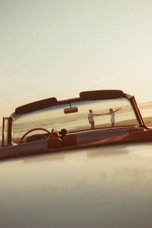 Couple standing on a beach at sunset, viewed through the windshield of a vintage convertible car.