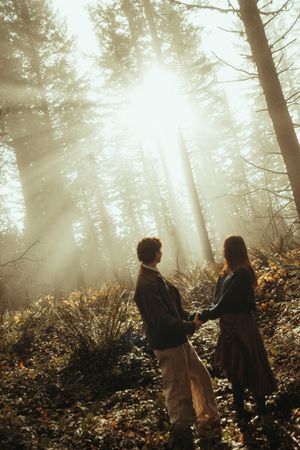 Two silhouettes stand in a misty forest with sunlight streaming through tall trees creating an ethereal atmosphere.