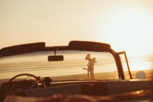 Couple embracing on a sunny beach at golden hour, viewed through a vintage convertible car windshield.