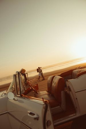 Couple embracing at sunset on a beach beside a classic white convertible car with warm golden light.