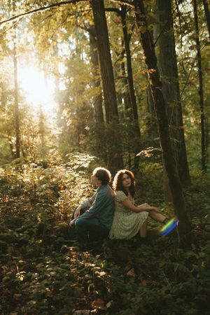 A couple sits together on a wooded hillside at sunset with sunlight streaming through autumn trees creating a magical glow.