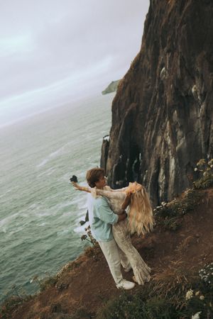 Two people in pastel clothing dance dramatically on the edge of a misty coastal cliff.