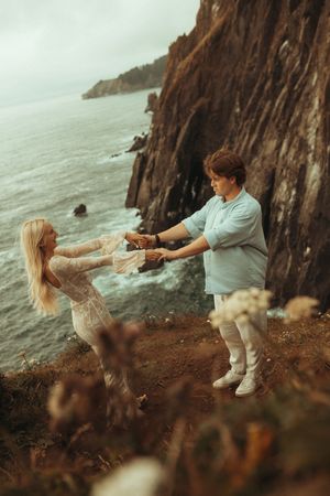 A couple playfully interacts on a dramatic coastal cliff overlooking the ocean on an overcast day.