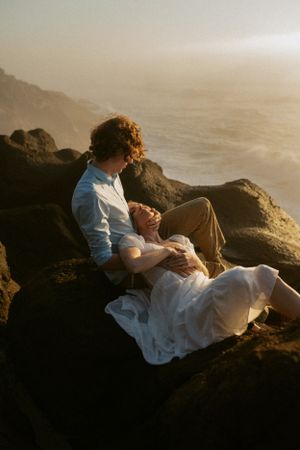 A couple in romantic embrace sits on coastal rocks at sunset with ocean waves and misty atmosphere in background.