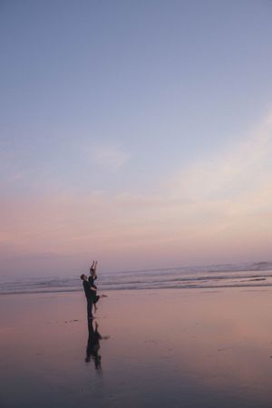 Couple silhouetted against a pastel pink and purple sunset sky on a reflective beach, one partner lifting the other.