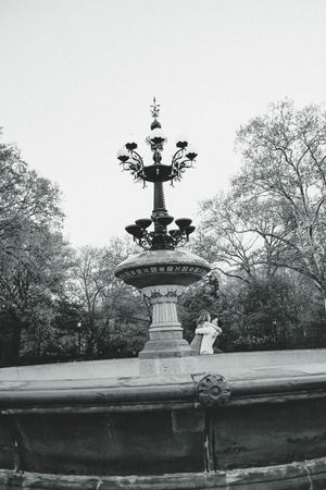 Black and white photo of an ornate vintage fountain with candelabra top in a park setting.