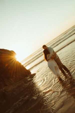 Silhouetted couple embraces on beach at sunset with golden sunlight streaming through.