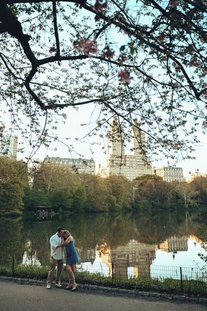 Couple embracing by a reflective lake in Central Park with cherry blossoms and NYC skyline at dusk.