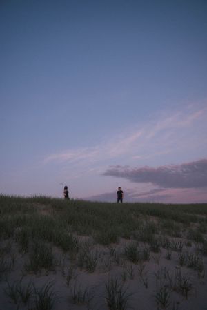 Two silhouetted figures stand on a grassy sand dune at dusk under a pastel blue and pink twilight sky.