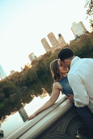 Couple sharing a romantic kiss on a bridge in Central Park with NYC skyline reflecting on the lake at golden hour.