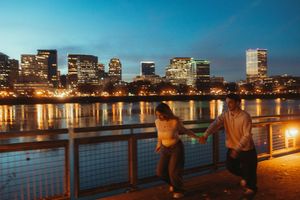 City skyline reflects on Charles River at dusk as people gather along illuminated waterfront promenade in Boston.