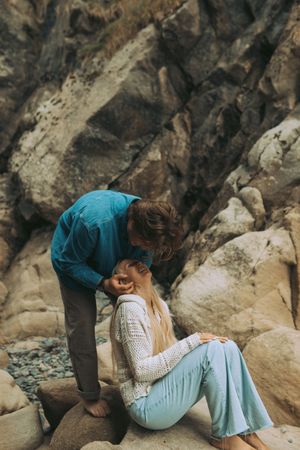 A couple shares an intimate moment on rocky coastal terrain wearing casual clothing in muted tones.