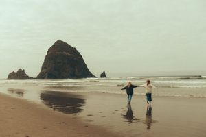 Two people walk along a misty beach with large rock formations visible in the distance on the Oregon coast.