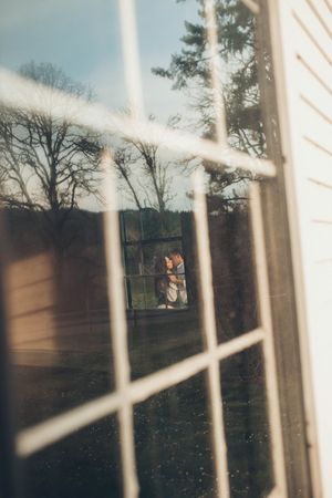Couple embracing reflected in a rustic window with bare trees and blue sky in the background.