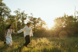Couple holding hands running through a sunlit meadow with golden hour light filtering through trees in a romantic outdoor setting.