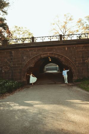Couple posing under a brick archway tunnel in Central Park, woman in flowing dress, man in light blue outfit.