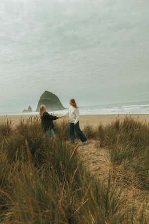 Two people walk through tall beach grass on a cloudy day near the ocean with dramatic cliffs in the distance.