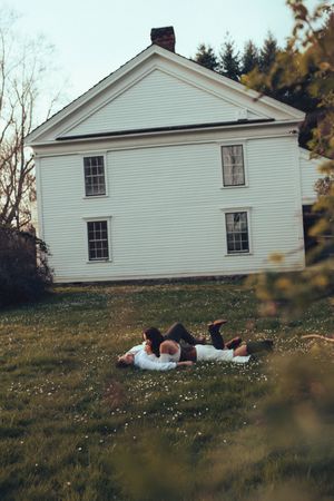 Couple lying on green grass in front of a classic white colonial farmhouse surrounded by trees.