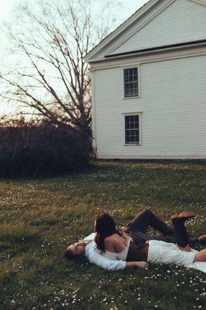 Couple lying on grass lawn beside a white colonial house with blooming wildflowers on a spring evening.
