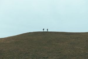 Two distant silhouettes walk along the ridge of a grassy hilltop against a bright sky.