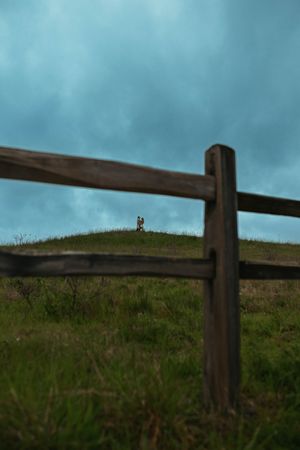 Wooden fence in foreground with couple standing on green hill under dramatic stormy blue sky.
