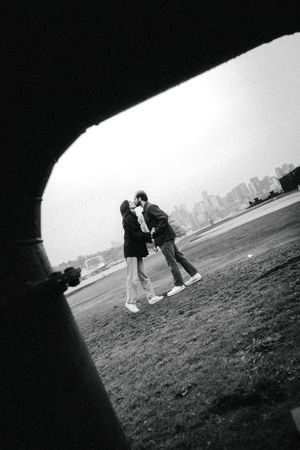Two figures stand in silhouette against a misty city skyline on a grassy hill in black and white photography.